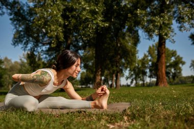 Side view of young caucasian sportswoman practicing yoga in half bound lotus forward fold pose in park. Girl wear sportswear with tattoos and closed eyes sit on fitness mat on lawn. Healthy lifestyle