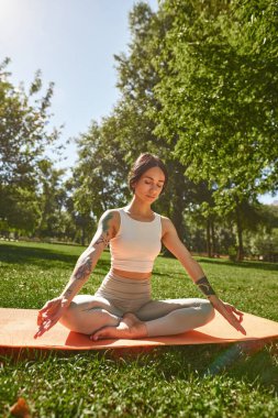 Young caucasian sportswoman practicing yoga in lotus pose and meditating in sunny park. Girl with tattoos and closed eyes wearing sportswear sitting on fitness mat on green meadow. Healthy lifestyle