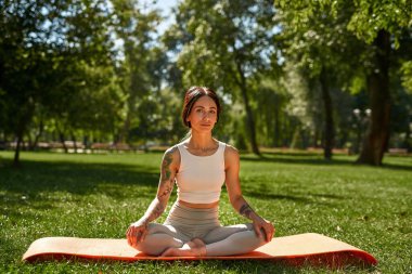 Young serious caucasian sportswoman practicing yoga and looking at camera in sunny park. Girl with tattoos wearing sportswear sitting on fitness mat on green lawn. Concept of healthy lifestyle