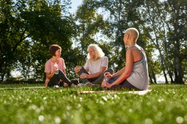 Woman looking at smiling elderly caucasian couple looking at each other in green sunny park. Friends resting and spending time together on fitness mats on meadow. Friendship. Healthy lifestyle