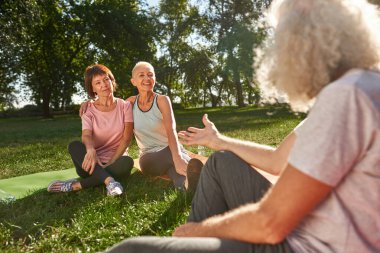 Partial grey hair man talking to senior caucasian women hugging in green sunny park. Friends resting and enjoying time on fitness mats on lawn. Friendship. Healthy lifestyle. Weekend and free time