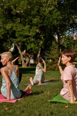 Female trainer showing and teaching mature caucasian women how practicing yoga in sunny park. Sportive female people sitting on fitness mats on green meadow. Concept of healthy lifestyle