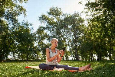 Focused european middle aged woman typing or chatting on smartphone in green sunny park. Sportive female pensioner wearing sportswear sitting on fitness mat on lawn. Concept of healthy lifestyle