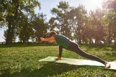Middle aged woman practicing yoga in park. Sportive female pensioner wearing sportswear standing on fitness mat on green lawn. Concept of healthy lifestyle. Sunny summer daytime