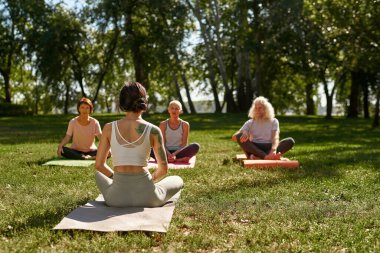 Back view of female trainer teaching elderly caucasian man and women how practicing yoga in sunny park. Sportive people sitting on fitness mats on green lawn. Concept of healthy lifestyle