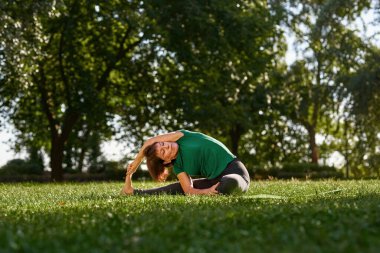 Caucasian mature woman practicing yoga in sunny park. Sportive female pensioner with closed eyes wearing sportswear sitting on fitness mat on green lawn. Concept of healthy lifestyle
