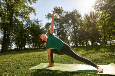 Caucasian senior woman practicing yoga in revolved side angel pose in sunny park. Sportive female pensioner wearing sportswear on fitness mat on green lawn. Concept of healthy lifestyle