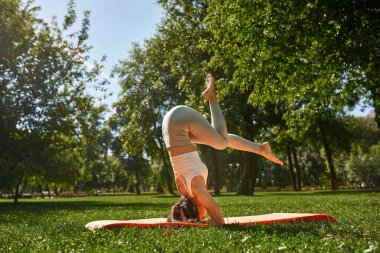 Side view of young caucasian athletic woman practicing yoga in half headstand pose in sunny park. Girl with tattoos wearing sportswear on fitness mat on green lawn. Concept of healthy lifestyle