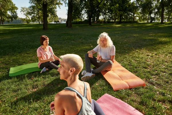 Elderly caucasian friends sitting, resting and drinking water after training on fitness mats on meadow. Sportive man and women. Friendship. Healthy lifestyle. Weekend and free time. Sunny green park