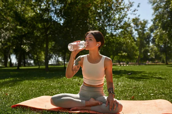 Young caucasian sportswoman drinking water from bottle on fitness mat on green lawn. Girl with tattoos wearing sportswear and wireless earphones resting after training in sunny park. Healthy lifestyle