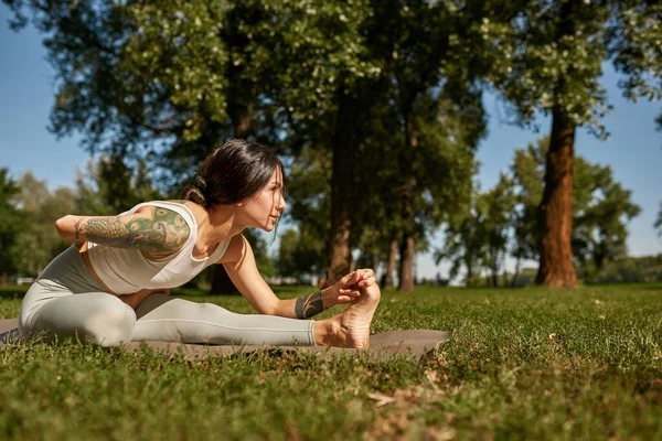 Side view of young caucasian sportswoman practicing yoga in half bound lotus forward fold pose in sunny park. Girl wearing sportswear with tattoos sitting on fitness mat on meadow. Healthy lifestyle