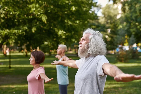 Selective focus of focused man with blurred friends practicing yoga on meadow. Partial side view of sportive elderly caucasian man and women. Friendship. Healthy lifestyle. Sunny green park