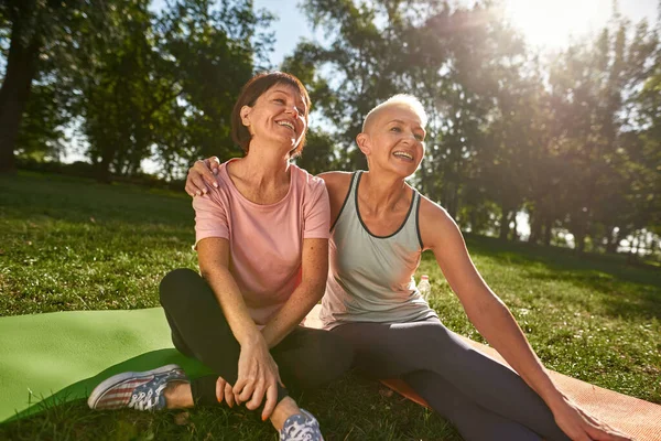 Pleased elderly caucasian women hugging and resting on fitness mats on meadow. Sportive female friends looking away. Friendship. Healthy lifestyle. Weekend and free time. Sunny green park