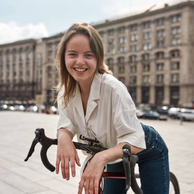 Smiling caucasian girl resting with her bicycle on blurred city square. Young pretty blonde woman looking at camera. Modern urban healthy female lifestyle. Entertainment, leisure and hobby. Cycling