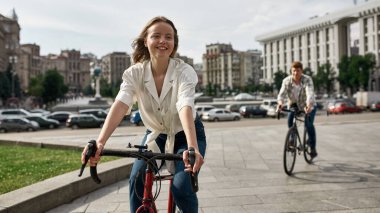 Focus on foreground of smiling caucasian girl and her boyfriend on background riding bicycles in city. Modern urban healthy lifestyle. Leisure and hobby. Biking. Young couple spending time together
