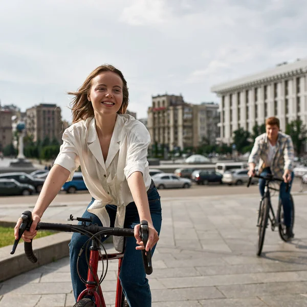 Focus on foreground of caucasian girl looking away while riding bicycles with her blurred boyfriend on background in city. Modern urban healthy lifestyle. Biking. Young couple spending time together