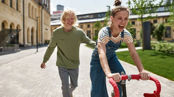 Guy running behind his excited girlfriend with open mouth riding bicycle in city. Modern urban healthy teenager lifestyle. Biking. Teen relationship. Young caucasain couple enjoying time together