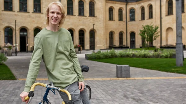 Young smiling blonde caucasian guy with his bicycle resting on city square and looking at camera. Modern urban healthy teenager lifestyle. Entertainment, leisure and hobby. Biking. Daytime