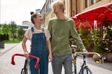 Young happy caucasian couple walking with bicycles in city. Modern urban teenager lifestyle. Leisure and hobby. Biking. Teen relationship. Guy and girl enjoying time together and looking at each other