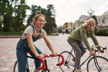 Young smiling caucasian couple riding bicycles and enjoying time together in city. Modern urban healthy teenager lifestyle. Entertainment, leisure and hobby. Biking. Blonde guy looking at girlfriend