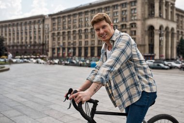 Young smiling caucasian man standing with his bicycle during riding on city square and looking at camera. Modern urban healthy male lifestyle. Entertainment, leisure and hobby. Biking. Daytime