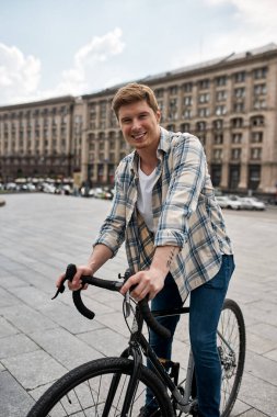 Young smiling caucasian man resting with his bicycle during riding on city square. Guy looking at camera. Modern urban healthy male lifestyle. Entertainment, leisure and hobby. Biking. Daytime