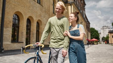 Young smiling caucasian couple talking and going with bicycle in city. Modern urban teenager lifestyle. Leisure and hobby. Biking. Teen relationship. Guy and girl spend time together. Warm sunny day