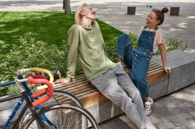 Young laughing caucasian couple sitting and resting on wooden bench on city square near bicycles. Modern urban teenager lifestyle. Teen relationship. Excited guy and girl enjoying time together