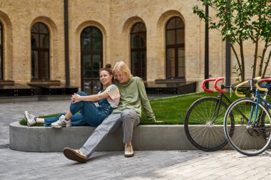 Young smiling caucasian couple sitting, resting and spending time together near near bicycles on city square. Modern urban teenager lifestyle. Teen relationship. Guy hugging his girlfriend. Sunny day