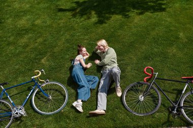 Top view of young smiling caucasian couple lying and looking at each other on green meadow with bicycles in sunny park. Modern teenager lifestyle. Rest and leisure. Guy and girl enjoying time together