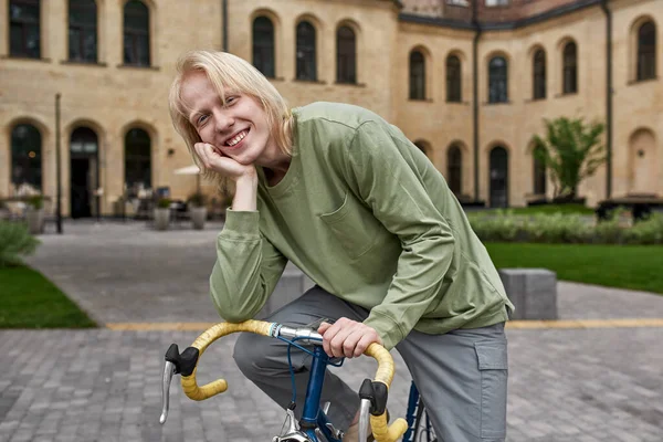 Young smiling blonde caucasian guy sitting on his bicycle on city square and looking at camera. Modern urban healthy teenager lifestyle. Entertainment, leisure and hobby. Biking. Daytime