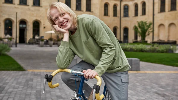 Young pleased blonde caucasian guy sitting on his bicycle on city square and looking at camera. Modern urban healthy teenager lifestyle. Entertainment, leisure and hobby. Cycling. Daytime