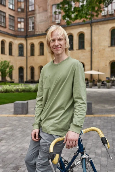 Young smiling blonde caucasian guy standing with his bicycle on city square and looking at camera. Modern urban healthy teenager lifestyle. Entertainment, leisure and hobby. Biking. Daytime