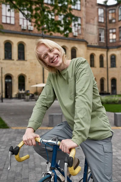 Young joyful blonde caucasian guy sitting on his bicycle on city square and looking at camera. Modern urban healthy teenager lifestyle. Entertainment, leisure and hobby. Biking. Daytime