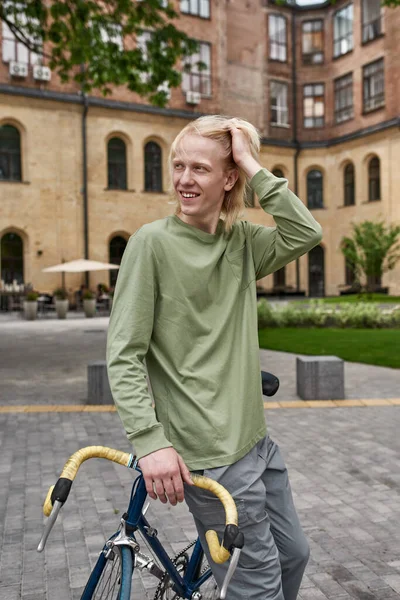Young smiling blonde caucasian guy standing with his bicycle on city square and looking away. Modern urban healthy teenager lifestyle. Entertainment, leisure and hobby. Biking. Daytime
