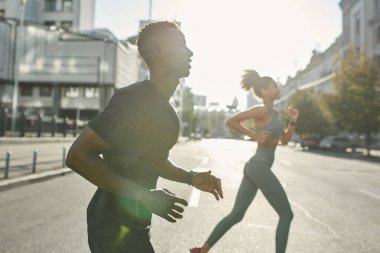 Focus on foreground of athletic sportsman running with blurred sportswoman on city street. Young slim black couple wearing sportswear. Concept of modern healthy lifestyle. Sunny day