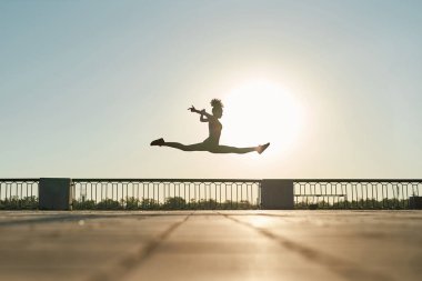 Side view of young black sportswoman jumping during running on sunny background in city. Beautiful curly slim girl wearing sportswear. Concept of modern healthy lifestyle. Warm sunny day