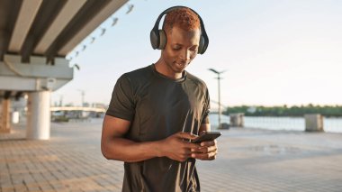 Young focused black sportsman using and watching smartphone on blurred city area. Athletic man wearing sportswear and headphones. Concept of modern healthy lifestyle. Sunny daytime