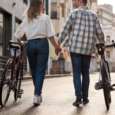 Back view of couple holding hands and walking with bicycles on city street. Modern urban healthy lifestyle. Entertainment, leisure and hobby. Relationship. Caucasian man and woman enjoy time together