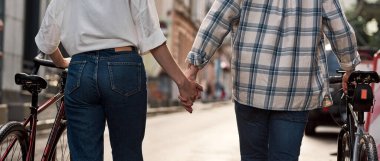 Cropped back view of couple holding hands and walking with bicycles on city street. Modern urban healthy lifestyle. Entertainment, leisure and hobby. Relationship. Man and girl spending time together