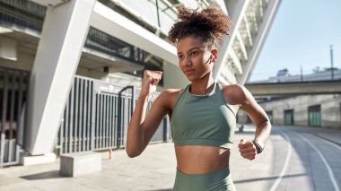 Young concentrated black sportswoman running or jogging near big stadium. Beautiful slim curly girl wearing sportswear and earphones. Concept of modern healthy lifestyle. Sunny day