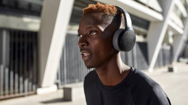 Cropped image of young serious black sportsman running or jogging near blurred big stadium. Handsome athletic guy wearing sportswear and headphones. Concept of modern healthy lifestyle. Warm sunny day