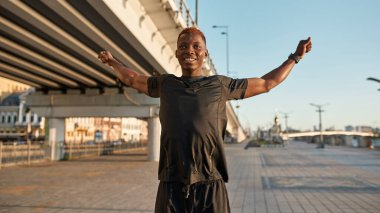 Young smiling black sportsman stretching on blurred city area. Slim athletic man wearing sportswear and looking away. Concept of modern healthy male lifestyle. Sunny daytime