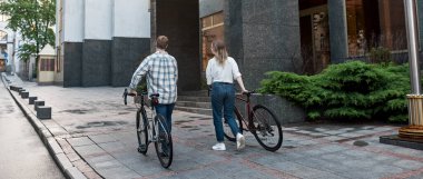 Back view of young couple walking with bicycles on city street. Modern urban healthy lifestyle. Entertainment, leisure and hobby. Biking. Man and girl speding time together. Daytime