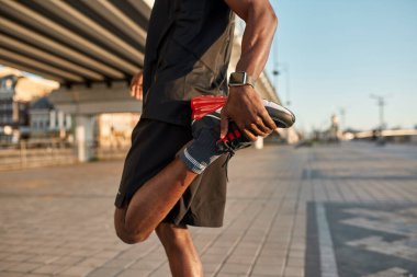 Partial down side view of black sportsman warming up leg on blurred city area. Slim athletic man wearing sportswear. Concept of modern healthy male lifestyle. Sunny daytime