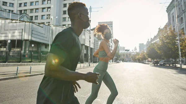 Young black sports couple running on city street. Attractive girl and athletic man wearing sportswear and medical masks. modern healthy lifestyle. Health protection during COVID-19 epidemic. Sunny day