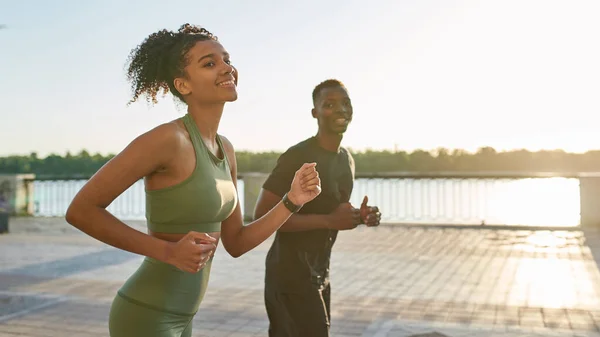 Young smiling black sports couple running on river promenade in city. Slim woman and athletic man wearing sportswear looking away. Concept of modern healthy lifestyle. Warm sunny day