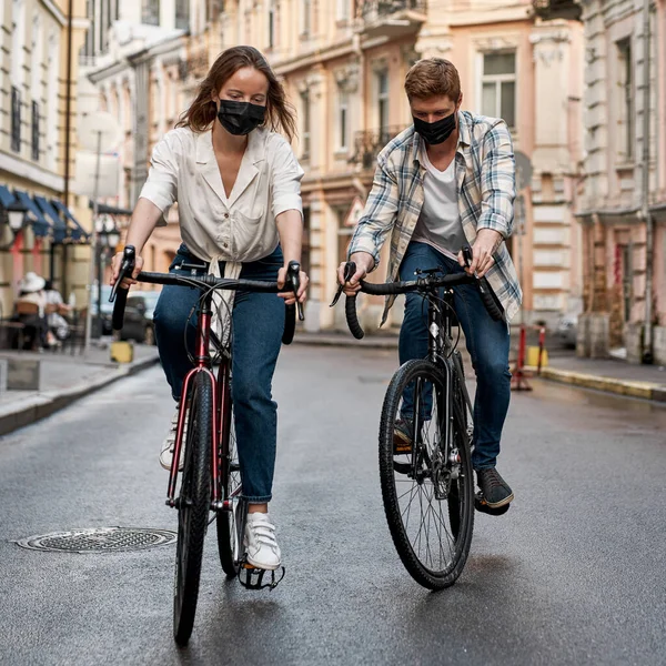 Young caucasian couple wear medical masks ride bicycles on street of historical city. Health protection during Coronavirus pandemic. Modern urban lifestyle. Entertainment, leisure and hobby. Biking