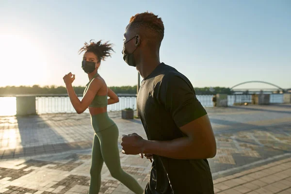 Young black sports couple running on river promenade in sunny city. Girl and athletic man wearing sportswear and medical masks. Modern healthy lifestyle. Health protection during COVID-19 epidemic