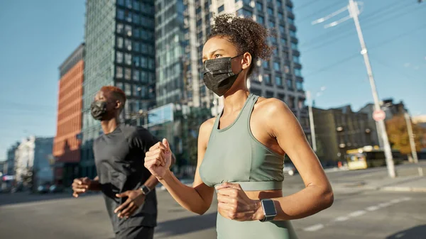 Young black sports couple running on city street. Beautiful girl and athletic man wear sportswear and medical masks. Modern healthy lifestyle. Health protection during Coronavirus pandemic. Sunny day